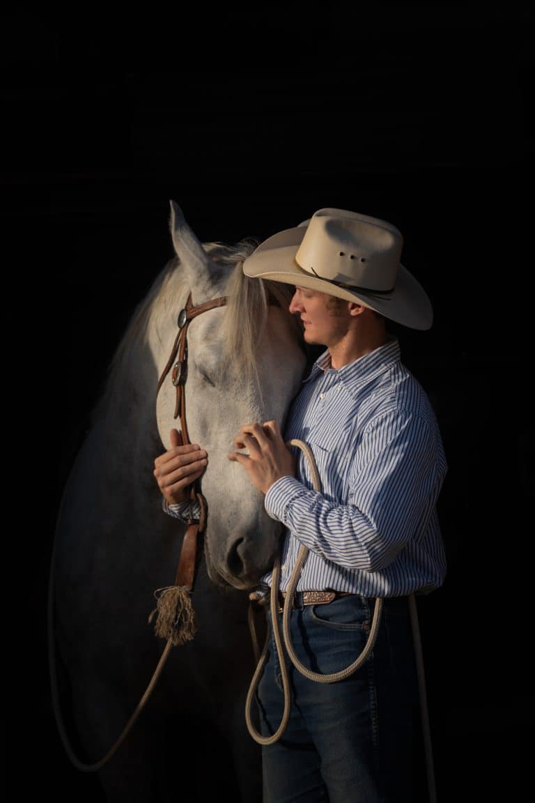 Horse trainer in a cowboy hat standing closely beside a horse against a dark background