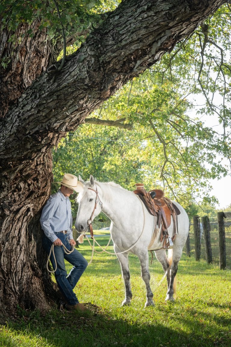Horse trainer in a cowboy hat leaning against a large tree beside a white horse in Kentucky