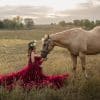Teen equestrian in a burgundy dress sitting in a field and looking up at her horse