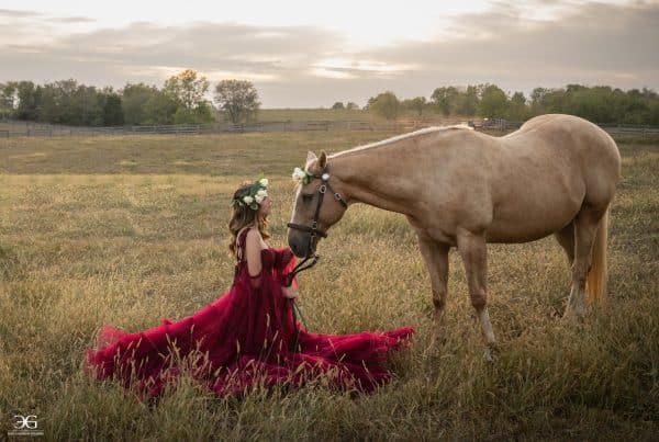 Teen equestrian in a burgundy dress sitting in a field and looking up at her horse