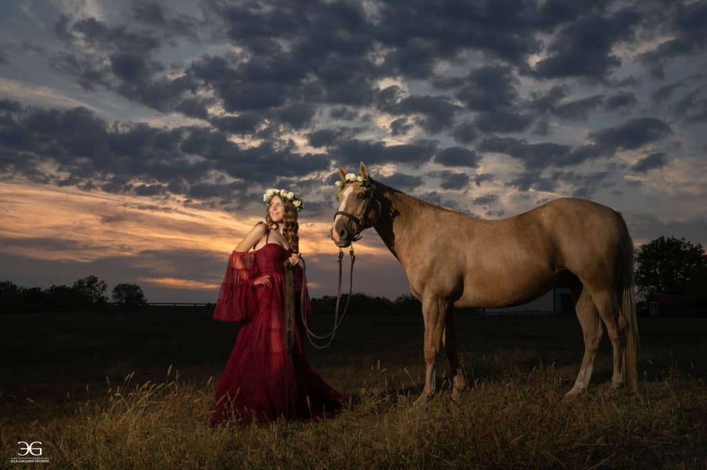 Teen girl in a burgundy gown standing beside her horse beneath a dramatic Kentucky sky