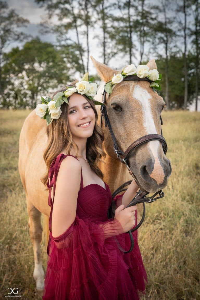 Teen girl in a burgundy dress and flower crown standing closely beside her horse