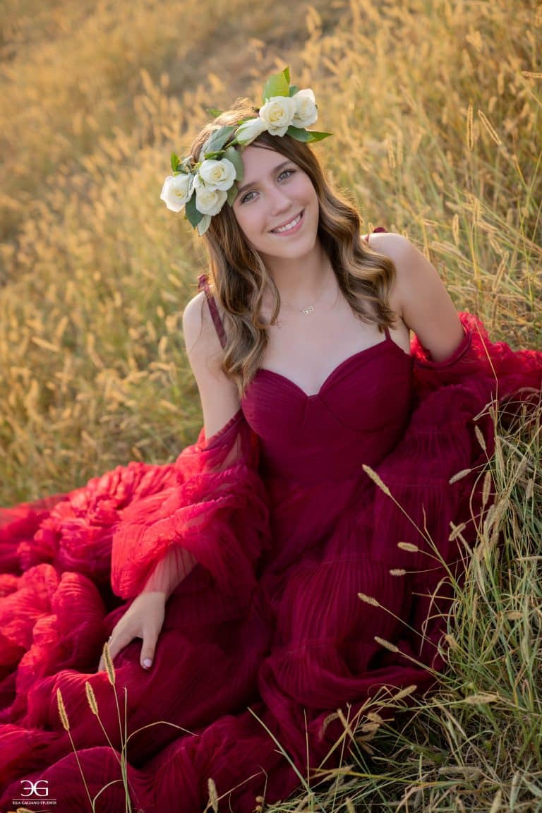 Teen equestrian in a burgundy gown and white flower crown sitting in a field